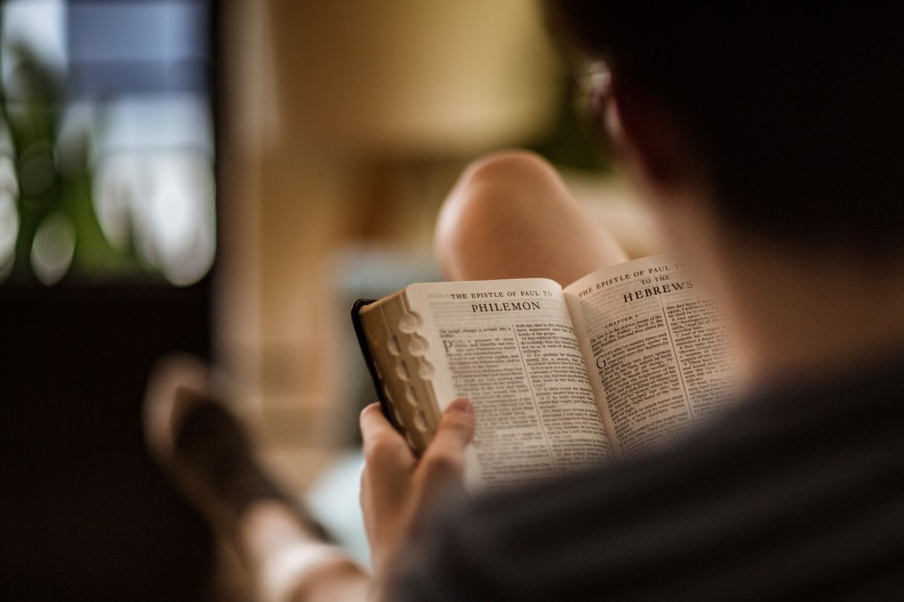 Un bărbat stând pe pat studiază Biblia sfântă cu paginile deschise la epistola lui Pavel către Filimon.